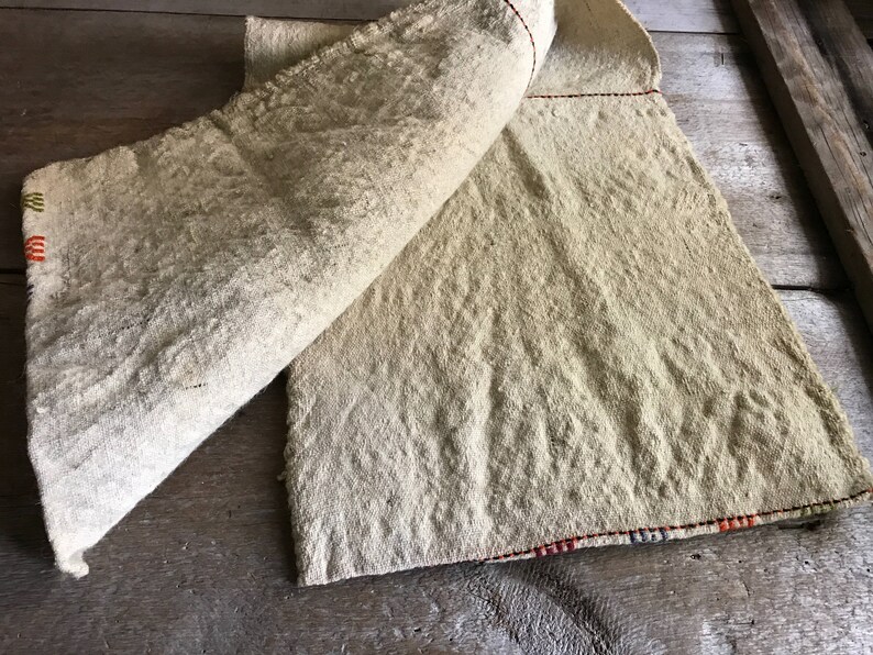 May include: A beige linen fabric with a red and orange striped border. The fabric is folded in half and has a rough, textured surface.