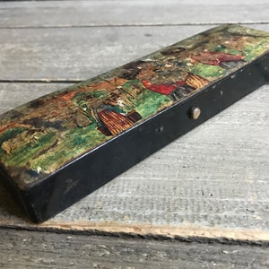 May include: A vintage black metal box with a hinged lid. The lid is decorated with a colorful painted scene of people walking in a park. The box has a small metal latch.