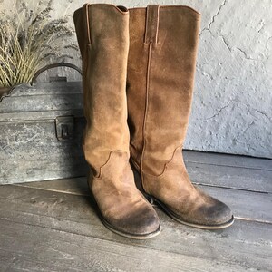 May include: A pair of brown suede knee-high boots. The boots have a worn, distressed appearance with a slight heel and a classic Western-inspired design. The boots are displayed on a wooden surface.