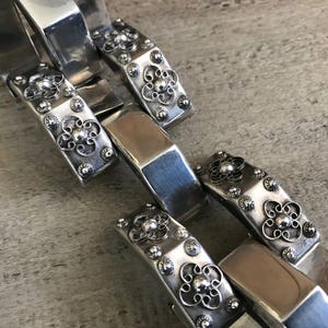 May include: A silver chain bracelet with intricate floral designs and small round accents. Each link is a rectangular shape with a raised floral pattern.