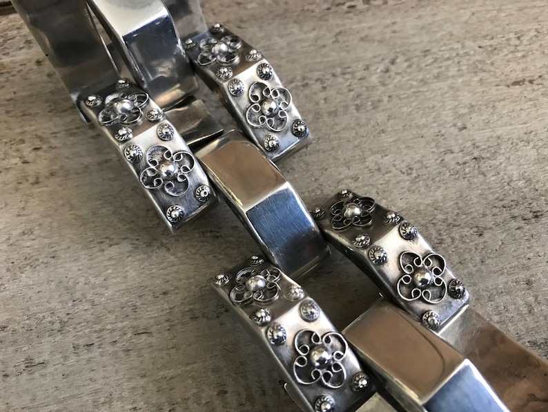 May include: A silver chain bracelet with intricate floral designs and small round accents. Each link is a rectangular shape with a raised floral pattern.