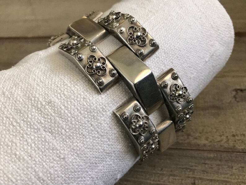 May include: A silver bracelet with a chain link design and intricate floral details. Each link is decorated with a small, raised flower.