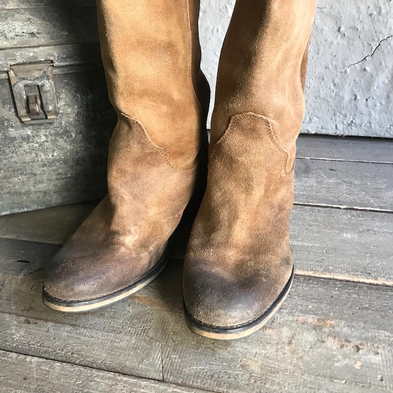 May include: A pair of tan suede boots with a worn appearance. The boots have a mid-calf height and a rounded toe. The soles are a light color with a dark border. The boots are placed on a weathered wooden surface.