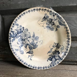 Könnte beinhalten: Ein runder Keramikteller in cremeweißer Grundfarbe mit einem dunkelblauen Blumenmuster. Das Design umfasst eine durchgehende Bordüre aus kleinen Blüten sowie detaillierte botanische Illustrationen von Clematis-Ranken.