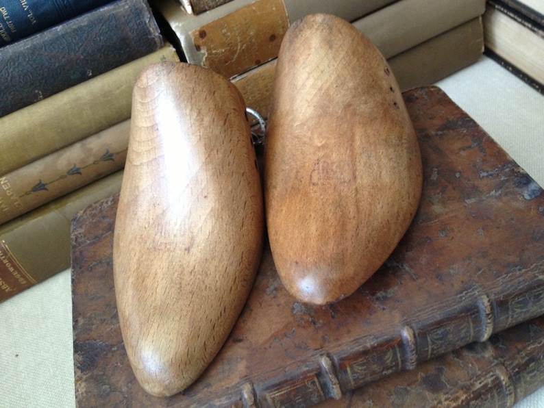 May include: Two wooden shoe trees, one on top of the other, with a light brown finish. The shoe trees are on top of a stack of old books.