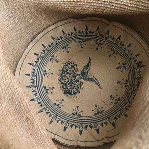 K&ouml;nnte beinhalten: Das Innere eines Strohhutes mit einem blauen und wei&szlig;en Blumendesign. Das Design zeigt einen stilisierten Vogel mit einer Blume im Schnabel.