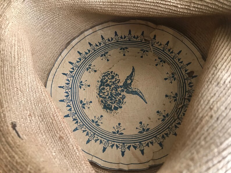 K&ouml;nnte beinhalten: Das Innere eines Strohhutes mit einem blauen und wei&szlig;en Blumendesign. Das Design zeigt einen stilisierten Vogel mit einer Blume im Schnabel.