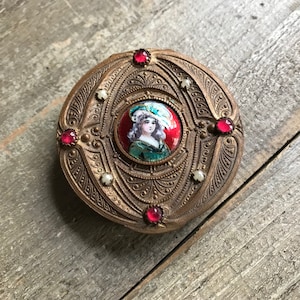 May include: A round, ornate, gold-toned metal compact with a red and white enamel portrait of a woman in a blue and white hat. The compact is decorated with red and white gemstones.