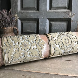 May include: A section of a decorative, antique wooden column with intricate floral and geometric carvings. The column features a light beige and white color scheme with a weathered, distressed appearance. The column is resting on a wooden surface.