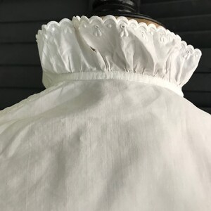 Pu&ograve; includere: Una camicia bianca in stile vintage con colletto a balze. Il colletto presenta un delicato bordo smerlato con piccoli dettagli ricamati. Il tessuto della camicia appare leggero e leggermente strutturato, suggerendo una miscela di lino o cotone.