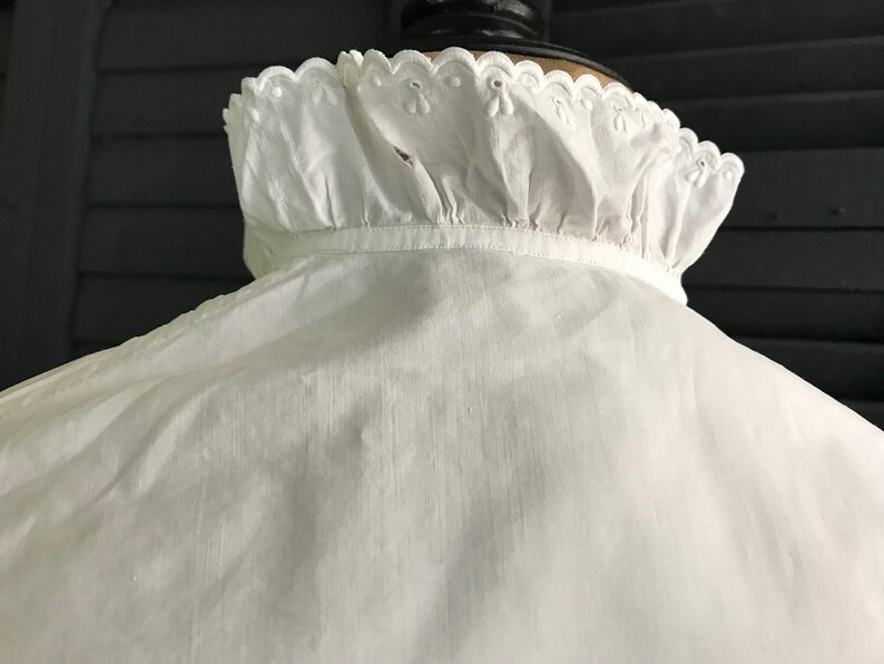 Pu&ograve; includere: Una camicia bianca in stile vintage con colletto a balze. Il colletto presenta un delicato bordo smerlato con piccoli dettagli ricamati. Il tessuto della camicia appare leggero e leggermente strutturato, suggerendo una miscela di lino o cotone.