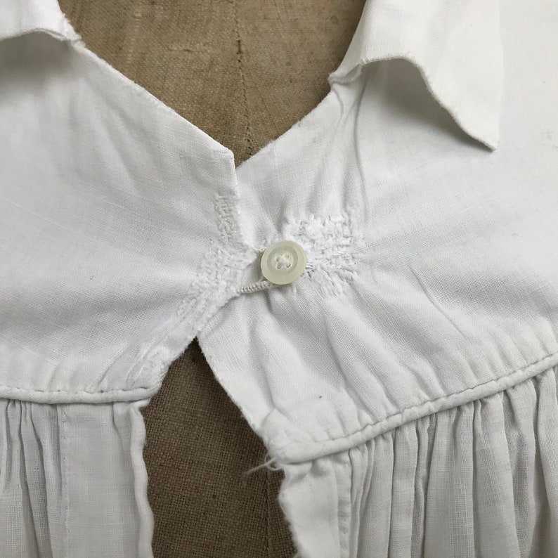 May include: White cotton shirt with a button closure at the neck. The shirt has a gathered detail at the bottom.
