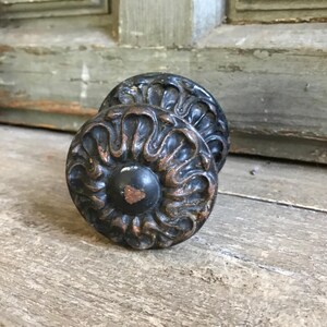 Puede incluir: Dos pomos de puerta redondos y antiguos con un acabado oscuro y desgastado. Los pomos tienen un diseño central circular con patrones ornamentados y ondulados en color bronce. El fondo es una superficie de madera envejecida.