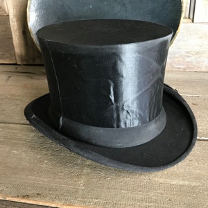 May include: A black top hat with a satin finish and a wide brim. The hat is sitting on a wooden surface.