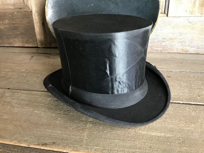 May include: A black top hat with a satin finish and a wide brim. The hat is sitting on a wooden surface.