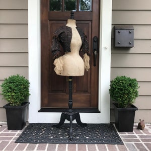 May include: A vintage dress form with a brown and black bodice on a black wooden stand. The dress form is in front of a wooden door with a black door handle. The dress form is in front of a house with a brown siding.