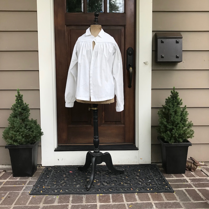 May include: A white, long-sleeved shirt with a gathered neckline and a button-up front. The shirt is displayed on a black mannequin stand against a wooden door.