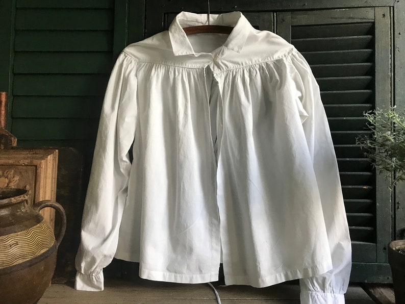 May include: A white, long-sleeved shirt with a gathered neckline and a tie closure. The shirt is made of a lightweight fabric and has a relaxed fit.