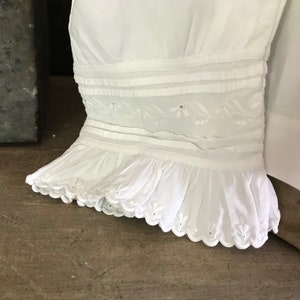 Pu&ograve; includere: Primo piano di una manica di un capo bianco. La manica presenta fasce orizzontali, ricami floreali e un orlo a balze con bordi smerlati. Il tessuto sembra essere cotone o un materiale leggero simile.