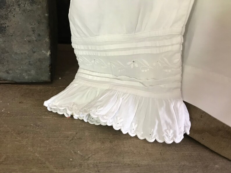 Pu&ograve; includere: Primo piano di una manica di un capo bianco. La manica presenta fasce orizzontali, ricami floreali e un orlo a balze con bordi smerlati. Il tessuto sembra essere cotone o un materiale leggero simile.