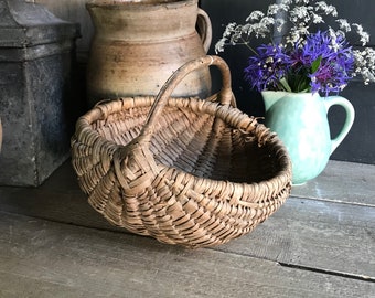 May include: A woven wicker basket with a handle, sitting on a wooden surface. The basket is brown and has a rounded shape. There is a clay pot and a pitcher with flowers in the background.