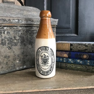 Puede incluir: Una botella de cerámica marrón y blanca con una etiqueta que dice "The Carlisle Old Brewery Co. Ltd. Extra Double Stout." La etiqueta presenta un escudo con un corazón y una corona.