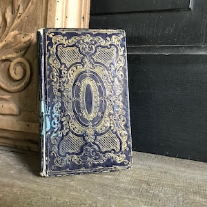 May include: A vintage book with a dark blue cover and gold ornate design. The cover features a central oval shape with a gold border.