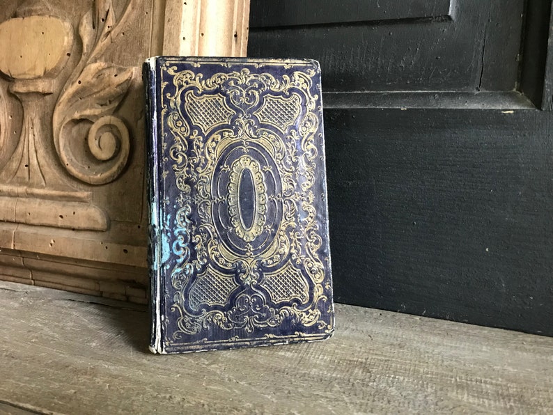 May include: A vintage book with a dark blue cover and gold ornate design. The cover features a central oval shape with a gold border.