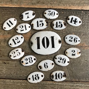 Op de afbeelding: Verzameling vintage witte ovale emaille huisnummerplaten met zwarte cijfers. Nummers zijn onder meer 2, 6, 7, 10, 11, 12, 13, 14, 15, 18, 21, 23, 26, 28, 29, 30 en 101. De platen zijn gerangschikt op een verweerd houten oppervlak.