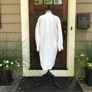 May include: A white long-sleeved shirt with a gathered neckline and long, wide sleeves. The shirt is displayed on a black mannequin stand against a brown wooden door.
