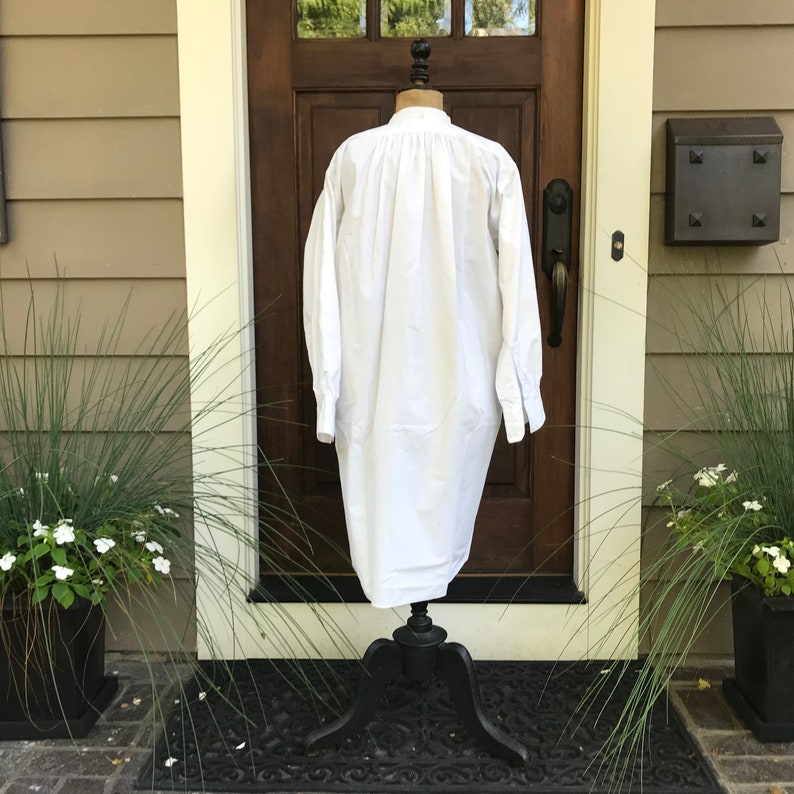 May include: A white long-sleeved shirt with a gathered neckline and long, wide sleeves. The shirt is displayed on a black mannequin stand against a brown wooden door.