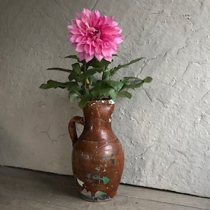 May include: A single pink dahlia flower in a brown ceramic jug with a green glaze. The jug has a handle and a rounded body. The flower is in bloom and has many petals.