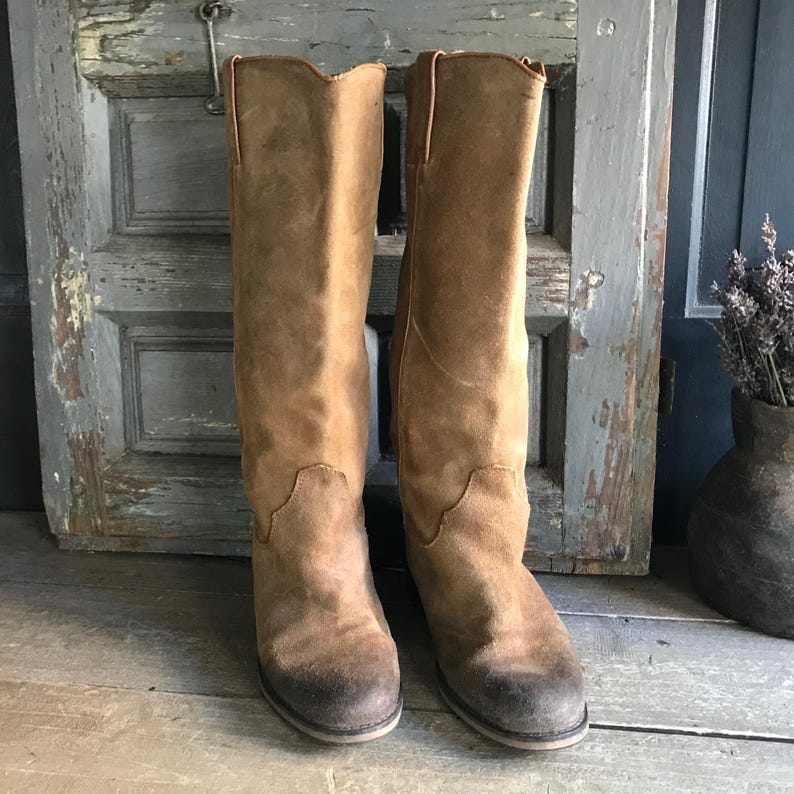 May include: A pair of tall, tan suede boots with a distressed appearance. The boots have a classic Western style with a notched top and a subtle heel. The worn look adds character to these knee-high boots, perfect for a vintage or bohemian aesthetic.