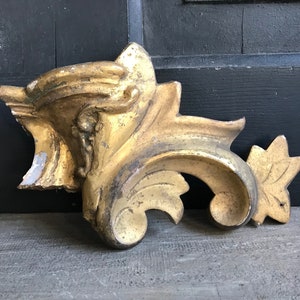 Könnte beinhalten: Antikes, goldfarbenes Fragment einer architektonischen Zierleiste. Das dekorative Stück weist ein stilisiertes Blatt- und Rollendesign mit sichtbaren Abnutzungserscheinungen auf, die auf das Alter hindeuten. Die Leiste ist wahrscheinlich aus Holz und ein dekoratives Element.
