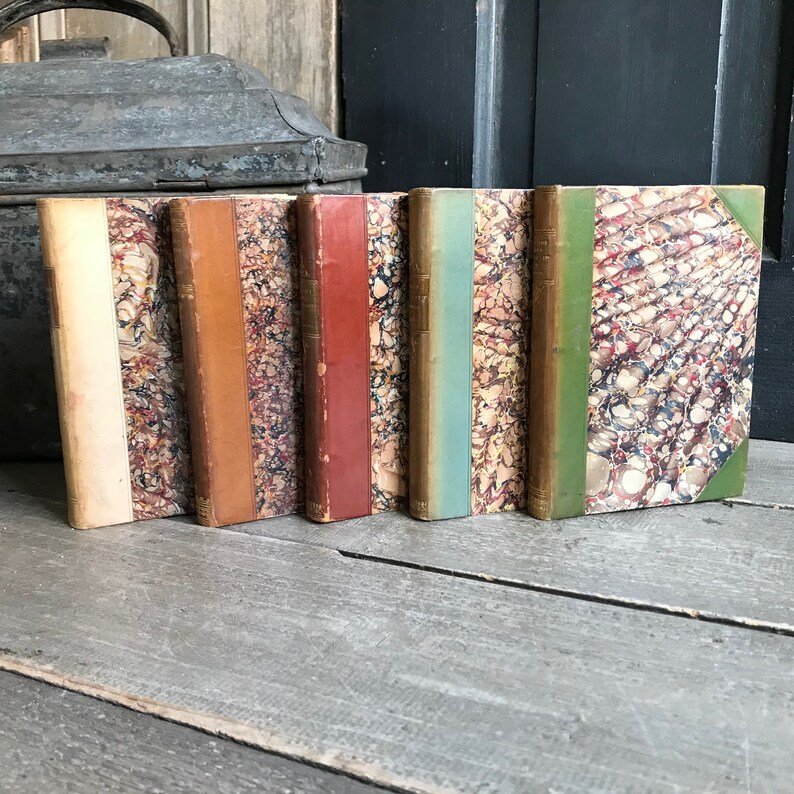 May include: A set of six antique books with marbled covers in various colors, including white, orange, red, blue, and green. The books are arranged in a row on a wooden surface.