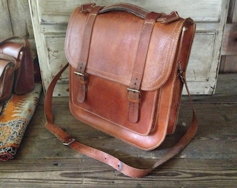 May include: A brown leather satchel with a long adjustable strap and two buckles on the front. The bag has a flap closure and a zippered pocket on the side.