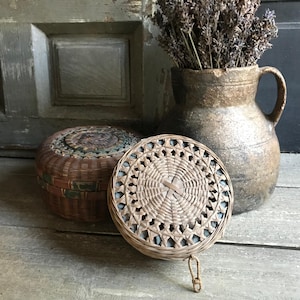 May include: Two woven baskets with lids and a rustic ceramic jug holding dried lavender. The baskets are brown and tan, with intricate woven patterns. The jug is a mottled brown with a handle. The items are arranged on a wooden surface.