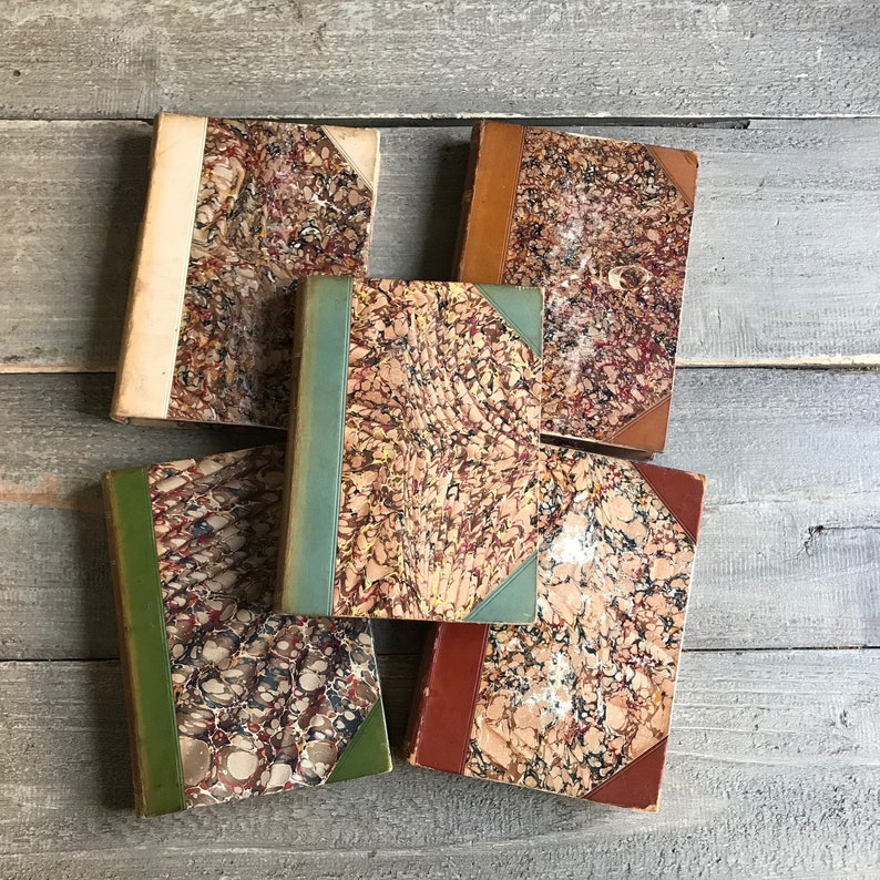 May include: Five antique books with marbled covers in shades of brown, green, and white. The books have a vintage aesthetic and are arranged in a stack.