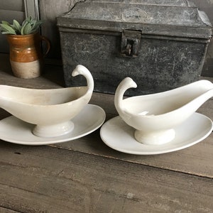 May include: Two white ceramic gravy boats with swan-shaped handles and matching saucers. The boats are slightly different in size and shape.
