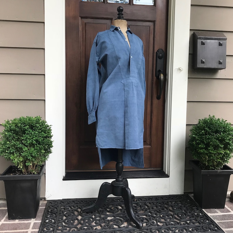 May include: A long-sleeved, light blue shirt with a high collar and a button-up front. The shirt is displayed on a black mannequin stand against a backdrop of a wooden door and a brick wall.