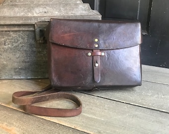 Pode incluir: Uma bolsa de couro marrom escuro com fecho de aba e uma longa alça de ombro. A bolsa apresenta rebites de latão e um fecho de tira de couro. Uma caixa de metal antiga está no fundo.