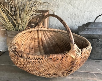May include: A large, woven, brown wicker basket with a single, curved wooden handle. The basket is empty and has a rustic, vintage look.