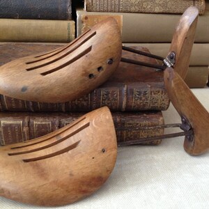 May include: Three antique wooden shoe stretchers with metal components. The shoe forms are light brown with several parallel slots. The stretchers are resting on a stack of old books with gold lettering.