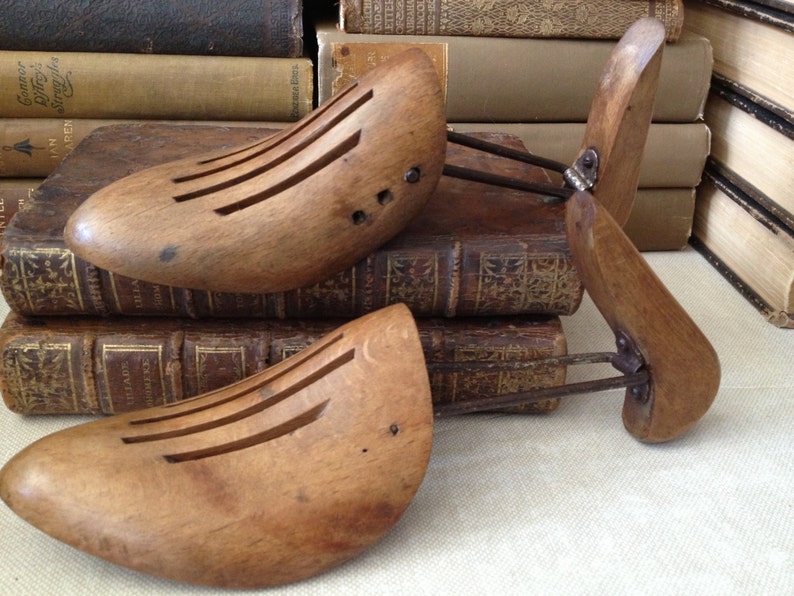 May include: Three antique wooden shoe stretchers with metal components. The shoe forms are light brown with several parallel slots. The stretchers are resting on a stack of old books with gold lettering.