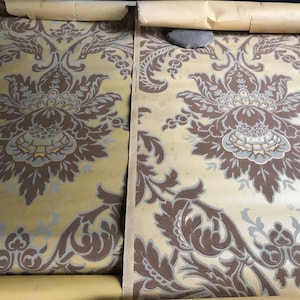 May include: Two rolls of vintage wallpaper featuring a repeating floral damask pattern. The design includes large, stylized flowers and leaves in shades of brown, gray, and light blue against a pale yellow background. The paper is rolled up.