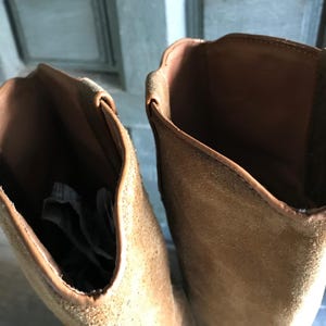 May include: Close-up of two brown suede boots. The boots have a tall shaft and a rounded top. The interior of the boots is a darker brown, and the exterior is a lighter tan color. The boots appear to be unworn.