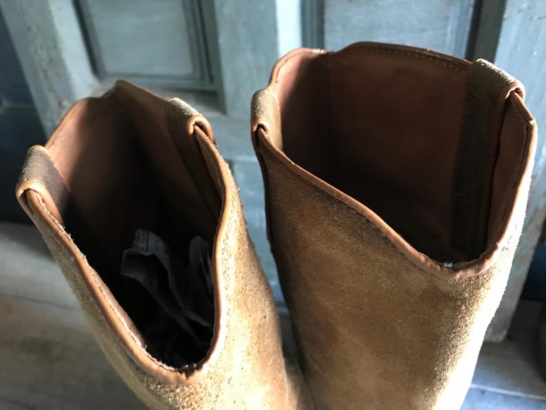 May include: Close-up of two brown suede boots. The boots have a tall shaft and a rounded top. The interior of the boots is a darker brown, and the exterior is a lighter tan color. The boots appear to be unworn.