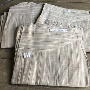 May include: Three folded, beige striped pants with white and brown stripes. Each pant has a tag that reads "MADE IN COTON". The pants are arranged on a wooden surface.