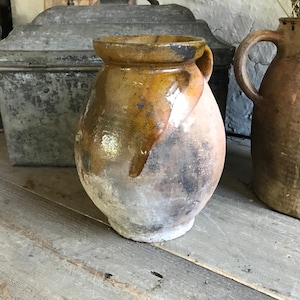 May include: A rustic, glazed ceramic pot with a handle. The pot is a mottled brown and yellow color with a white glaze. The pot is sitting on a wooden surface.