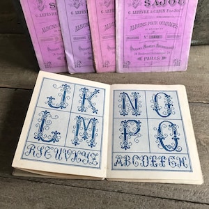 May include: Vintage French embroidery pattern books with pink covers and blue and white cross-stitch alphabet designs. The open book displays letters J, K, L, M, N, O, P, and Q. The books are titled "Albums Pour Ouvrages" and "Dessins Modeles Fournitures".